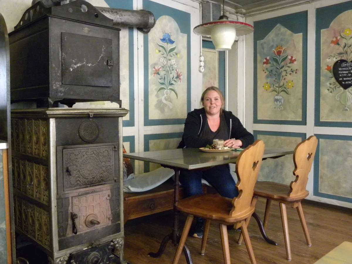 Eine Frau sitzt an einem Tisch. Die Wände erinnern an ein altes Gasthaus, daneben steht ein Holzofen.